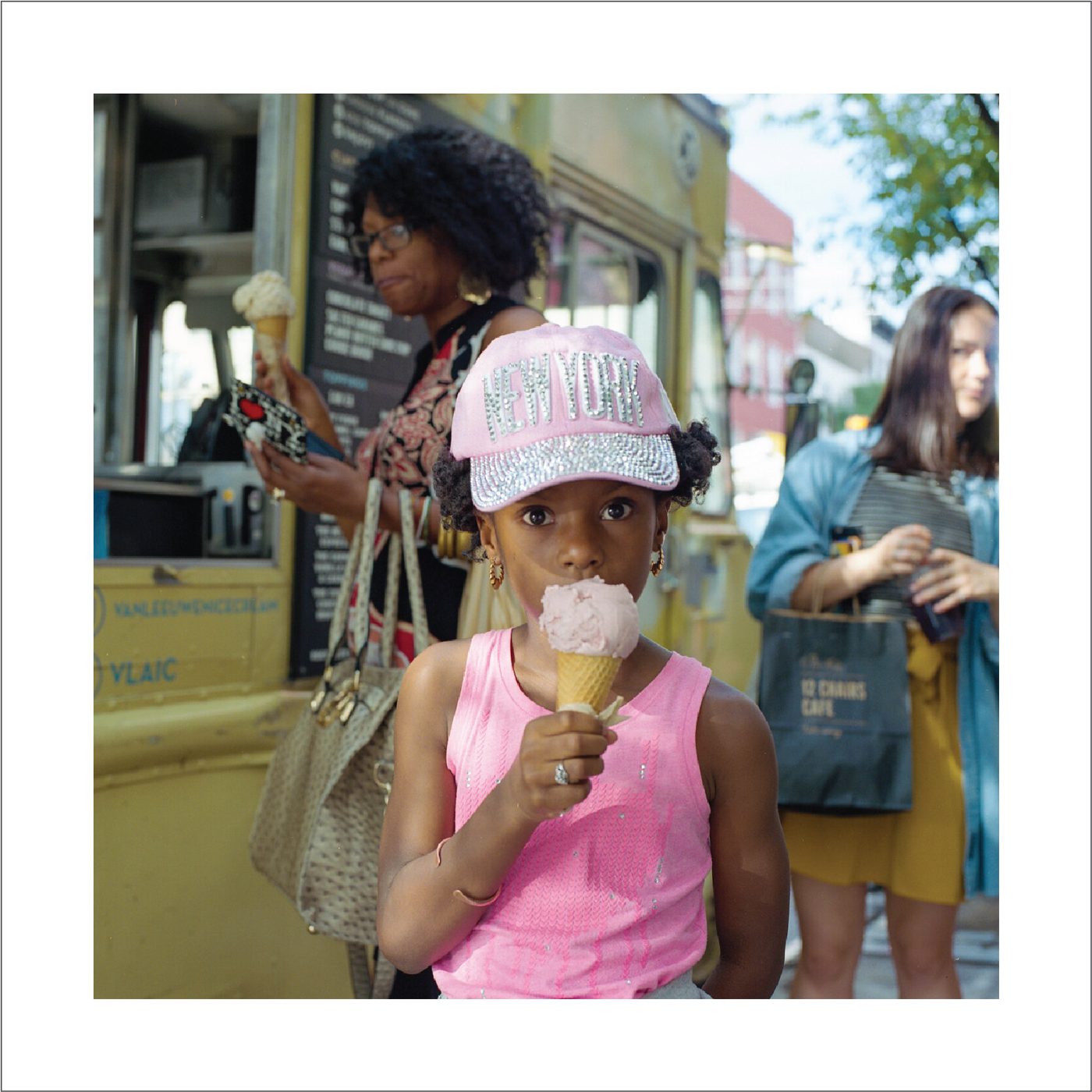 A young girl in a pink New York cap eats an ice cream cone on a Brooklyn sidewalk while adults wait behind her.